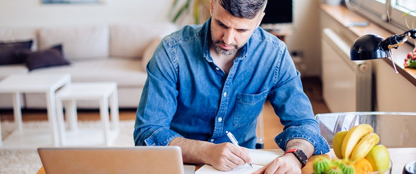 Junger Mann sitzt zuhause am Laptop und macht sich Notizen in einem Block
