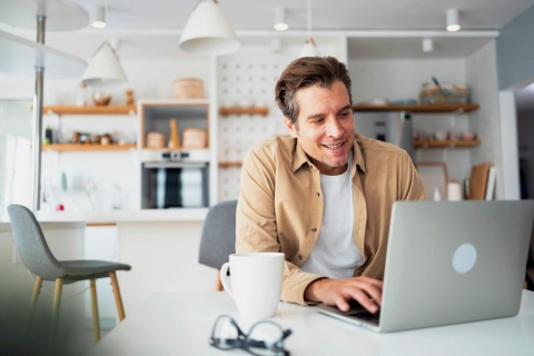 Mann in Küche sitzt vor dem Laptop und lacht